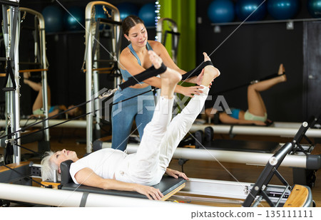 Coach helps elderly woman do leg exercises on reformer Coach helps elderly woman do leg exercises on reformer 135111111