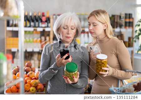 Girl and grandma scan QR code, take reading pickled olives label using phone camera in shop Girl and grandma scan QR code, take reading pickled olives label using phone camera in shop 135111284