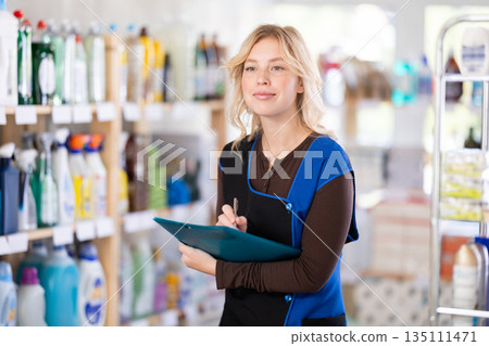 Beautiful diligent young girl saleswoman standing between shelves and doing inventory in hypermarket 135111471