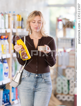 Girl housewife in household store examines goods, chooses liquid furniture polish Girl housewife in household store examines goods, chooses liquid furniture polish 135111507