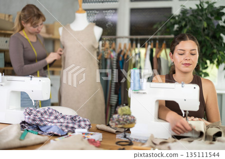 Fashion designer working at sewing machine in studio with teammate Fashion designer working at sewing machine in studio with teammate 135111544