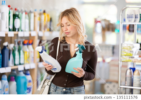 Attentive young girl choosing between two sprays standing between shelves in supermarket 135111576