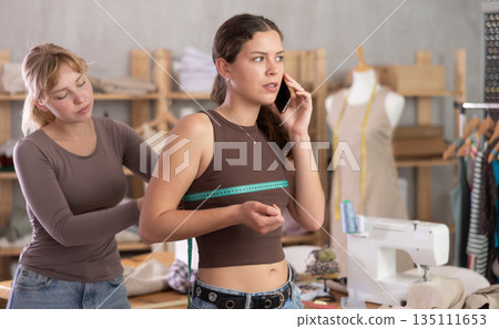 Young tailoress measuring female client speaking on mobile 135111653