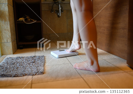 Young woman weighing herself in the bathroom, feet on the scale in front of the sink, slender 135112365