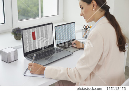 Female accountant reviewing spreadsheets on two laptops, taking notes in office 135112453