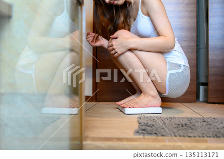 一位年輕女子在浴室裡稱體重。她身材苗條,很高興能減肥。 一位年輕女子在浴室裡稱體重。她身材苗條,很高興能減肥。 135113171
