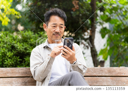 一名中年男子坐在長椅上操作智慧型手機 135113359
