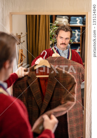 Man customer buying vintage jacket in second hand store, holding clothing and looking in mirror. Man customer buying vintage jacket in second hand store, holding clothing and looking in mirror. 135114840