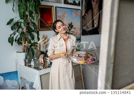 Pleased woman artist painting picture at workshop, looking at canvas, enjoying art hobby atmosphere. 135114841