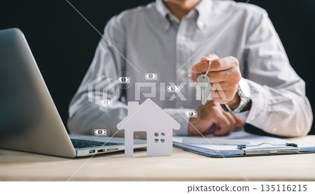 Real estate agent hand holds house key, desk features paper home model and property document, laptop screen shows mortgage loan for buying home, investors plan future ownership 135116215