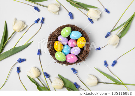 Spring background. Easter colored painted eggs in bird nest with white tulips and blue muscari on gray background with space for text and greetings. Easter and spring concept. Spring background. Easter colored painted eggs in bird nest with white tulips and blue muscari on gray background with space for text and greetings. Easter and spring concept. 135116998