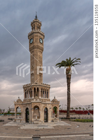 Iconic Konak Clock Tower and a tall palm tree in Konak Square, Izmir, Turkey, under a cloudy sky. Iconic Konak Clock Tower and a tall palm tree in Konak Square, Izmir, Turkey, under a cloudy sky. 135119358