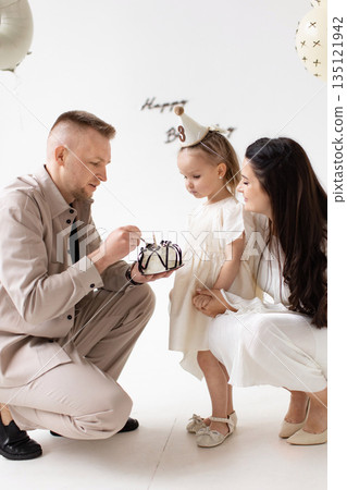A family celebrates a little girl's birthday with a cake and balloons in a bright, white room. A family celebrates a little girl's birthday with a cake and balloons in a bright, white room. 135121942