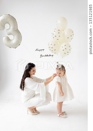 A mother helps her daughter with a birthday hat, surrounded by balloons and a 'Happy Birthday' message. 135121985