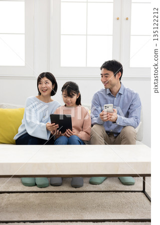 A family of three sitting on a sofa and smiling while operating a tablet and smartphone A family of three sitting on a sofa and smiling while operating a tablet and smartphone 135122212