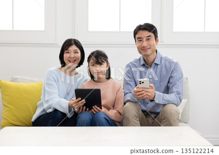 A family of three sitting on a sofa and smiling while operating a tablet and smartphone A family of three sitting on a sofa and smiling while operating a tablet and smartphone 135122214