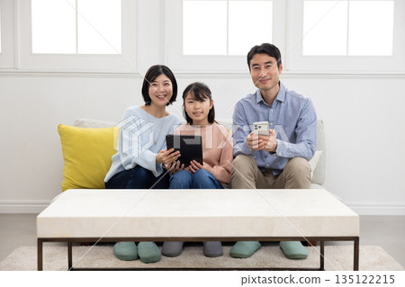 A family of three sitting on a sofa and smiling while operating a tablet and smartphone A family of three sitting on a sofa and smiling while operating a tablet and smartphone 135122215