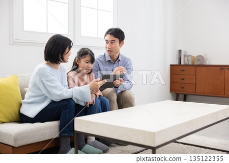 A family of three sitting on a sofa and smiling while operating a tablet and smartphone A family of three sitting on a sofa and smiling while operating a tablet and smartphone 135122355
