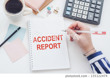 A hand with a pen writes text ACCIDENT REPORT in a notebook on a white desk. View from above. 135122474