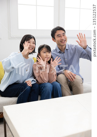 A family of three sitting on a sofa and waving with a smile 135122537
