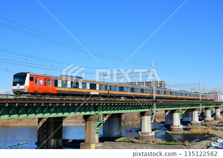 Under a blue sky, E233 series 0 blue 660 train crosses the Tama River, decorated to commemorate the 100th anniversary of the opening of the Itsukaichi Line. 135122562