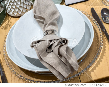 Close-up of a modern dining table set with white plates, a beige linen napkin, and a metallic napkin ring. The scene is softly lit, highlighting the elegant simplicity and cozy design of the tabletop Close-up of a modern dining table set with white plates, a beige linen napkin, and a metallic napkin ring. The scene is softly lit, highlighting the elegant simplicity and cozy design of the tabletop 135123212