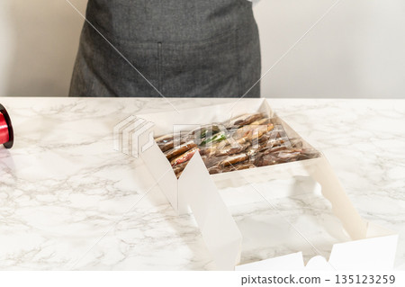 A person is carefully arranging wrapped Christmas cookies in a festive gift box. The box is filled with a variety of decorated cookies, perfect for gifting during the holiday season. This image A person is carefully arranging wrapped Christmas cookies in a festive gift box. The box is filled with a variety of decorated cookies, perfect for gifting during the holiday season. This image 135123259