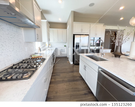 A side-angle view of a long modern kitchen featuring a built-in stove, white cabinetry, and stone countertops. Natural and ceiling light create a bright cooking space. A side-angle view of a long modern kitchen featuring a built-in stove, white cabinetry, and stone countertops. Natural and ceiling light create a bright cooking space. 135123260