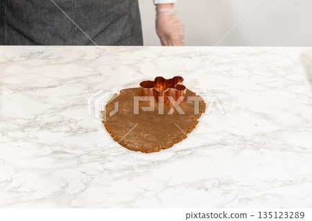Front-facing view of a gingerbread man cookie cutter pressed into spiced gingerbread dough on a marble countertop, emphasizing the shaping process. Front-facing view of a gingerbread man cookie cutter pressed into spiced gingerbread dough on a marble countertop, emphasizing the shaping process. 135123289