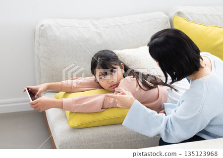An elementary school girl glaring at her mother while using a smartphone on the sofa An elementary school girl glaring at her mother while using a smartphone on the sofa 135123968