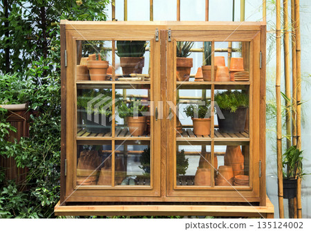 Rustic wooden garden cabinet with glass doors filled with terracotta pots and green plants. Perfect for small space gardening, home organization, and outdoor decor concept 135124002