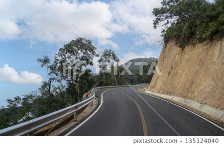 Scenic winding asphalt road on a lush green mountain under a beautiful blue sky with clouds. Perfect for travel, road trip, journey, and transportation infrastructure concept 135124040