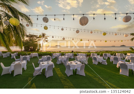 Luxury outdoor beach dinner setup at a tropical resort during golden hour sunset with lanterns. Perfect for destination wedding, event, and travel hospitality concepts 135124042