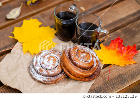 Two cups of hot coffee and cinnamon rolls on a wooden bench with autumn maple leaves in the park 135125129
