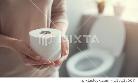 Person holding toilet paper roll near toilet bowl in bathroom with soft natural light Person holding toilet paper roll near toilet bowl in bathroom with soft natural light 135125507