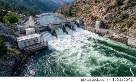 Hydroelectric dam releasing powerful water flow in rocky mountain river valley with lush green trees and clear sky 135125541
