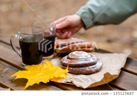 A hand reaches for a cup of coffee while sitting on a wooden bench outside among cinnamon buns and autumn maple leaves A hand reaches for a cup of coffee while sitting on a wooden bench outside among cinnamon buns and autumn maple leaves 135125687