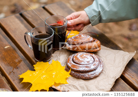 A hand reaches for a cup of coffee while sitting on a wooden bench outside among cinnamon buns and autumn maple leaves A hand reaches for a cup of coffee while sitting on a wooden bench outside among cinnamon buns and autumn maple leaves 135125688