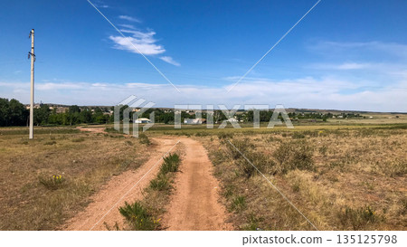 Dusty dirt road leading through a peaceful rural village with scattered houses and lush green trees under a clear blue sky. 135125798