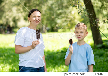 Mother and son eating chocolate ice cream on a stick in summer park 135126028