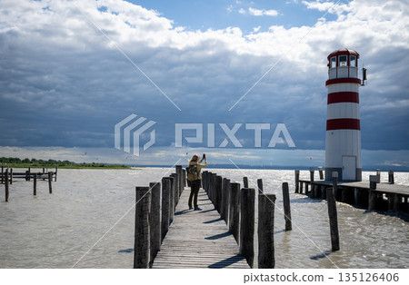 Traveler photographing Podersdorf Lighthouse Austria on windy pier by lake under dramatic storm clouds Traveler photographing Podersdorf Lighthouse Austria on windy pier by lake under dramatic storm clouds 135126406