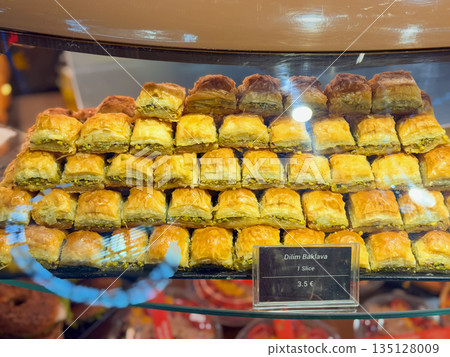 Freshly baked baklava slices are stacked neatly behind glass display in pastry shop, golden pastry and pistachio filling close up. Sweet food baklava, traditional dessert, symbol of sweet indulgence Freshly baked baklava slices are stacked neatly behind glass display in pastry shop, golden pastry and pistachio filling close up. Sweet food baklava, traditional dessert, symbol of sweet indulgence 135128009