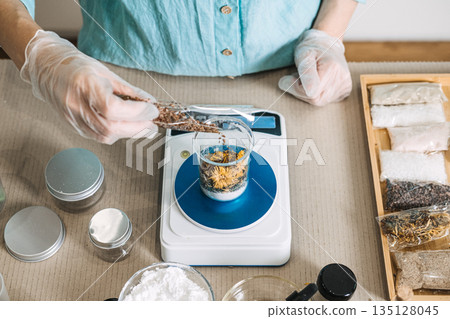 Hands in gloves pour brown seeds into glass beaker on digital scale near wooden tray with pouches. Cosmetic formulation, active ingredients, apothecary workshop, botanical science. 135128045