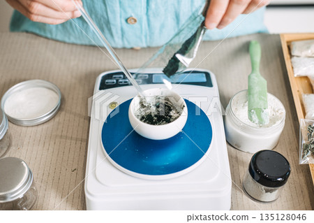 Hands pour dark powder from packet into small ceramic bowl on digital scale. Clean beauty standards, non-toxic formulation, cosmetic research and development, sustainable beauty lab. 135128046