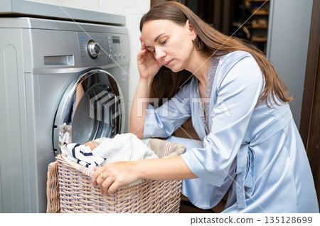 Tired housewife sitting by laundry basket with dirty clothes in bathroom 135128699