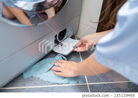 Woman cleaning washing machine drain filter through service hatch Woman cleaning washing machine drain filter through service hatch 135129468