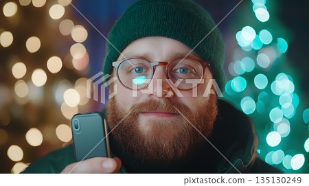 Young man in beanie holds smartphone, smiles against festive holiday lights background Young man in beanie holds smartphone, smiles against festive holiday lights background 135130249