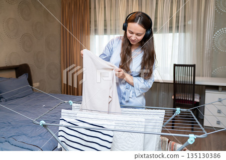Cheerful housewife listening to music with headphones while hanging laundry on drying rack Cheerful housewife listening to music with headphones while hanging laundry on drying rack 135130386
