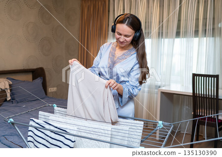 Cheerful housewife listening to music with headphones while hanging laundry on drying rack Cheerful housewife listening to music with headphones while hanging laundry on drying rack 135130390