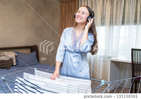 Cheerful housewife listening to music with headphones while hanging laundry on drying rack Cheerful housewife listening to music with headphones while hanging laundry on drying rack 135130391